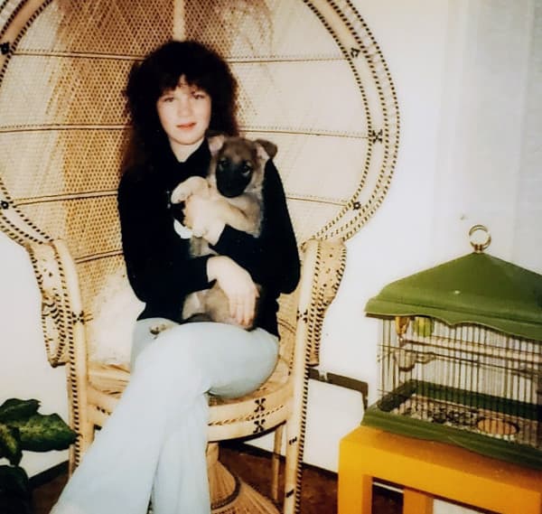The owner of Crewel Intentions as a teenager in the late 1970s, sitting in a wicker peacock chair holding a puppy, with a green birdcage beside her